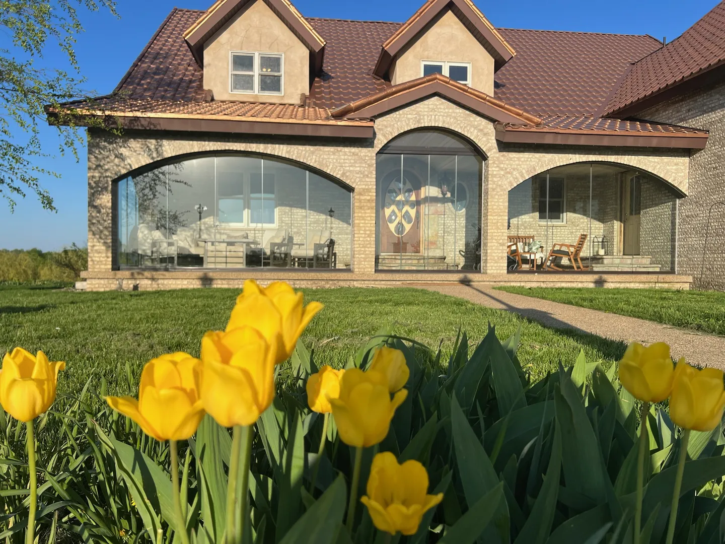 Stone house with arched porch openings enclosed in frameless glass, tulips in front