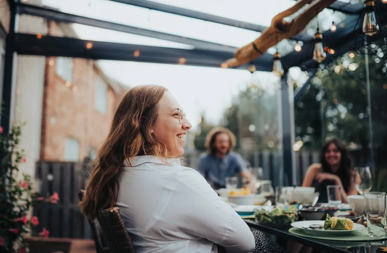 Friends laughing around a dinner table inside a glass patio enclosure