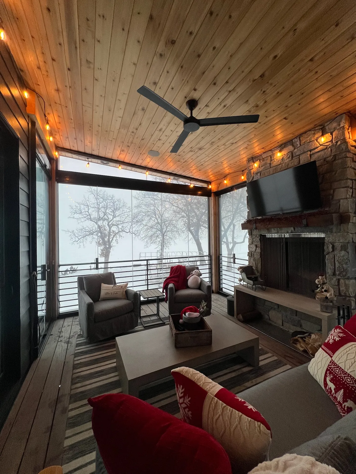 Armchairs and a stone fireplace inside a glazed porch, snow and fog beyond the glass